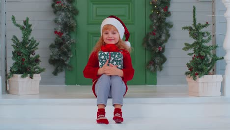 niño niño sonriente alegre sentado en el porche de la casa decorada sosteniendo una caja de navidad