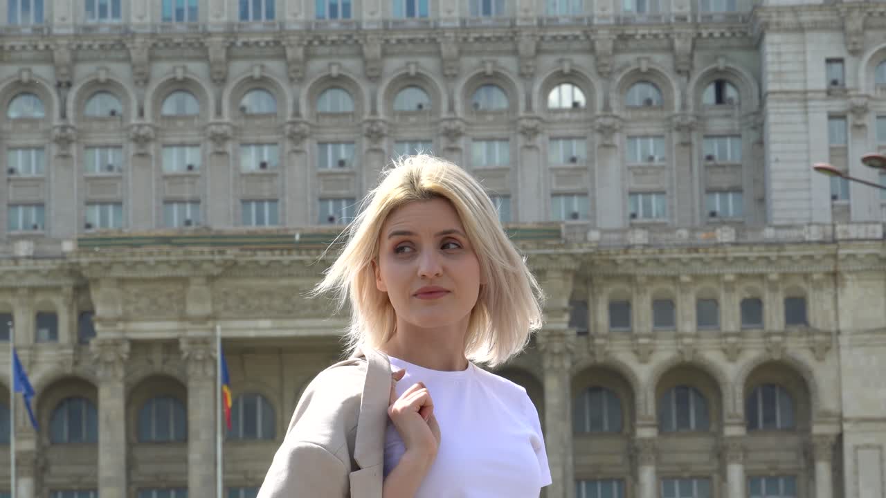 ⁇  티셔츠를 입은 젊은 금발 소녀가 누군가를 기다리고 있습니다. 배경에는 루마니아 부카레스트의 의회 궁전이 있습니다.