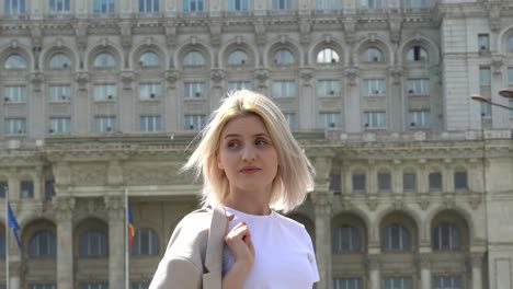 Young-Blonde-Girl-In-A-White-T-Shirt-Waiting-For-Someone,-Palace-Of-The-Parliament-In-The-Background,-Bucharest-Romania