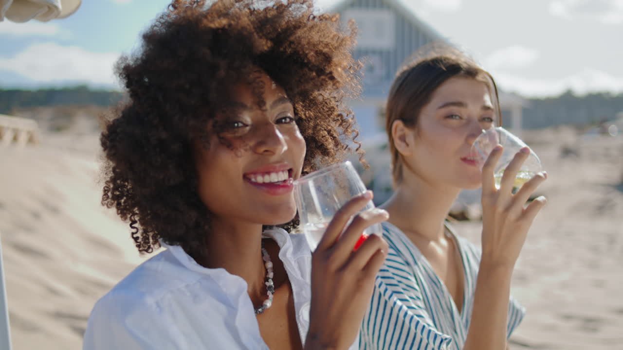 Closeup girls clinking glasses on summer beach picnic. Happy curly woman posing