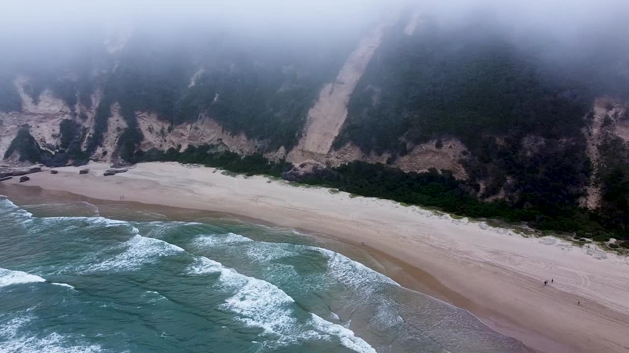 toma aérea de drones flotando debajo de la niebla de nubes bajas que se elevan hacia las dunas de arena que rodean la hermosa playa aislada en sedgefield, cabo occidental, sudáfrica