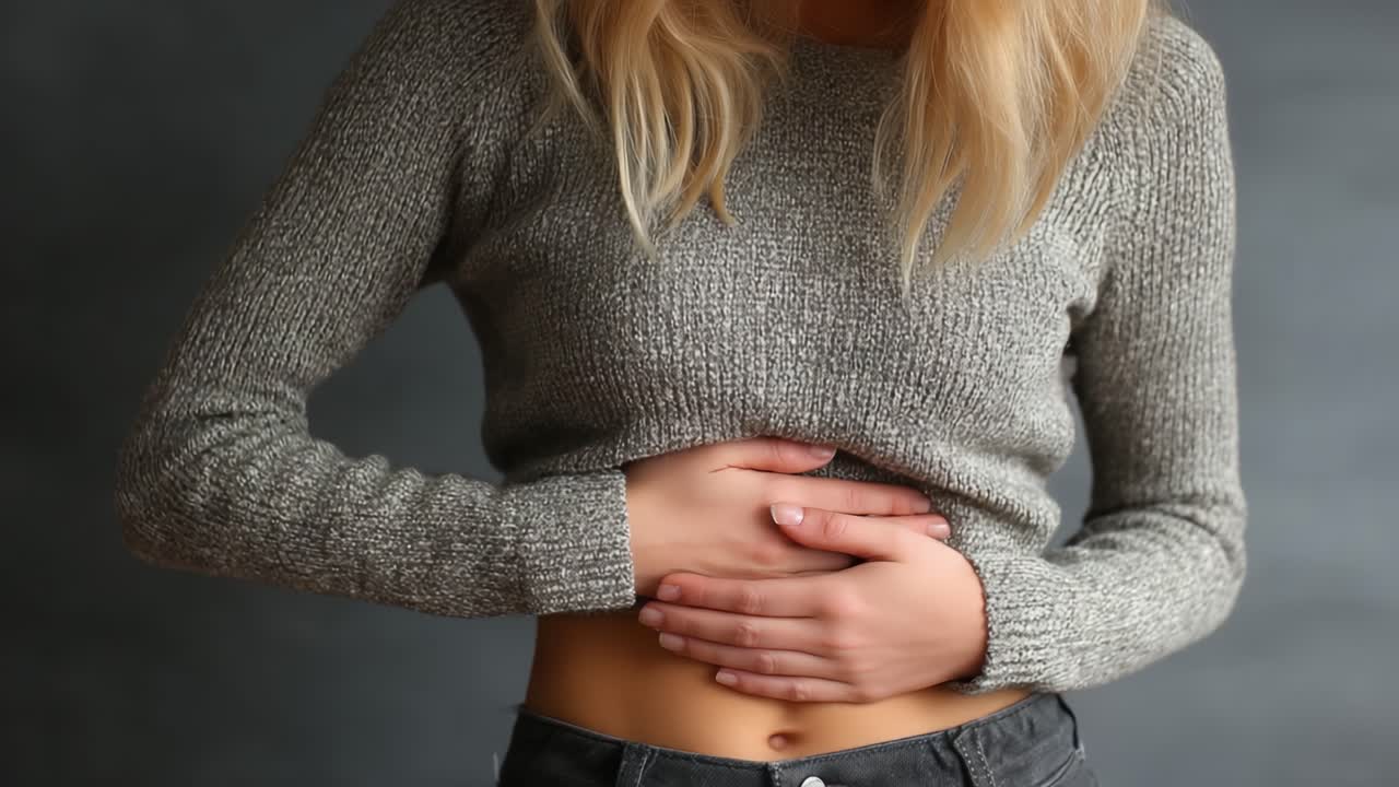 Una giovane donna con un maglione confortevole si tiene lo stomaco, indicando disagio o dolore, mentre si trova di fronte a uno sfondo grigio neutro con luce naturale che evidenzia la sua esperienza.