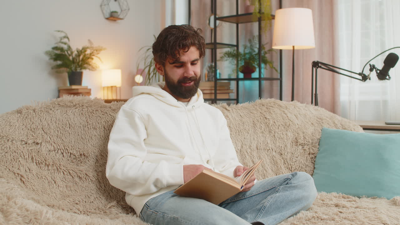 Indian man reading interesting book turning pages smiling enjoying literature sitting on sofa