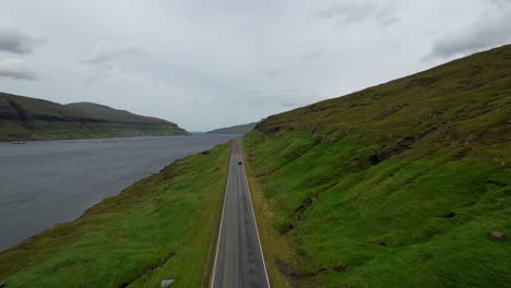 Drone-losing-track-of-car-driving-along-straight-road-on-green-hillside,-Faroe-Islands