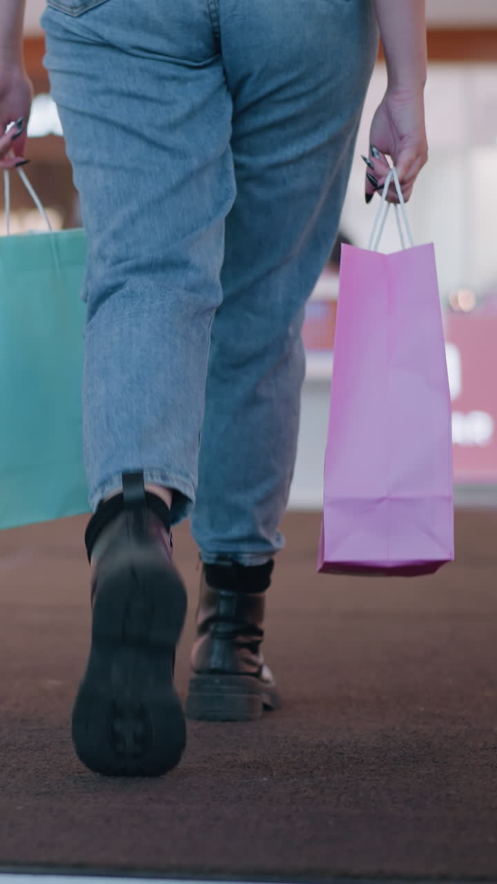 진즈와 부츠를 입은 사람이 움직이는 에스컬레이터에서 분홍색과 민트 쇼핑 가방을 들고 배경에 보케 조명과 함께 나가는 아래쪽 몸의 근접한 모습