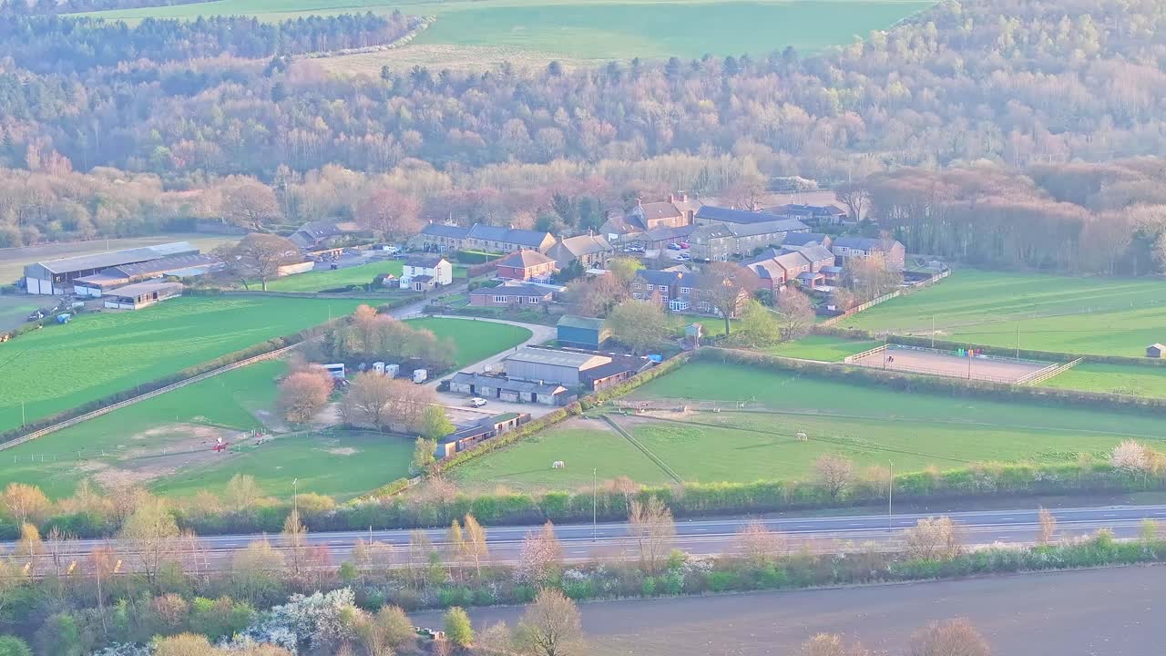 Static aerial drone shot overlooking a peaceful rural village with farm buildings, green fields, and an equestrian arena bordered by woodland in Wombwell, South Yorkshire, England