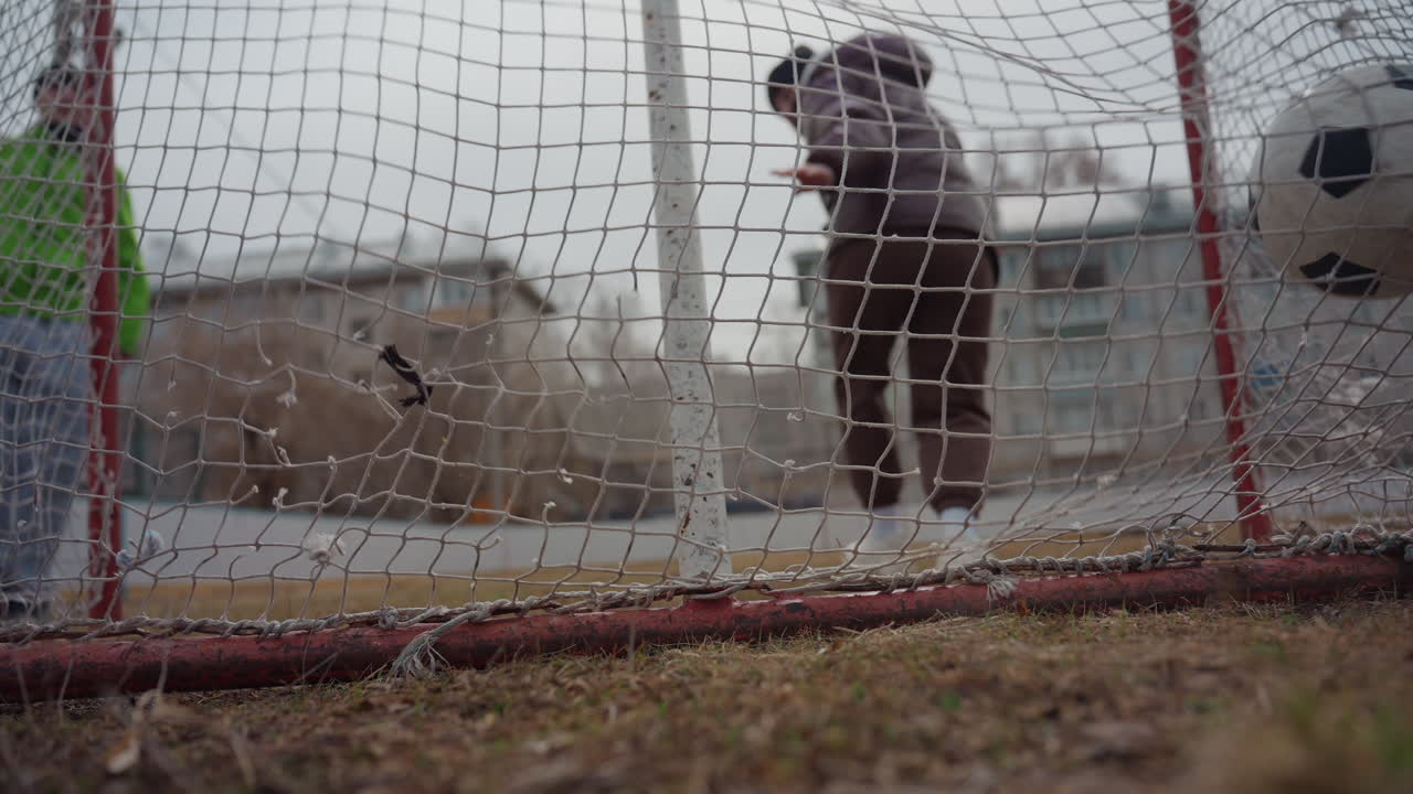 성공적인 세이브 시도의 로우 앵글 뷰, 녹색 옷을 입고 다가오는 성인과 아이 스트라이커의 실루엣, 흐린 하늘을 배경으로 그물에 닿는 공, 투박한 도시 놀이터의 긴장감과 역동적인 스포츠 동작