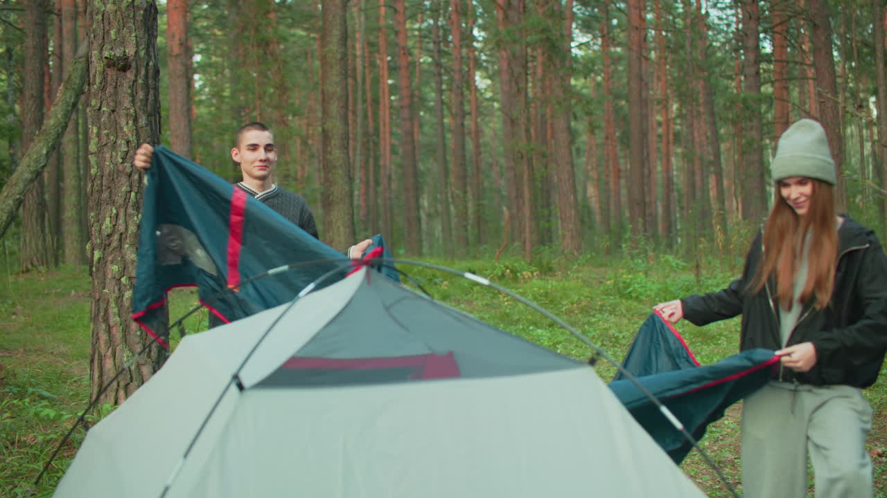 Couple works together to cover camping tent with outer sheet in forest clearing, both smiling and adjusting material while surrounded by tall trees and natural greenery