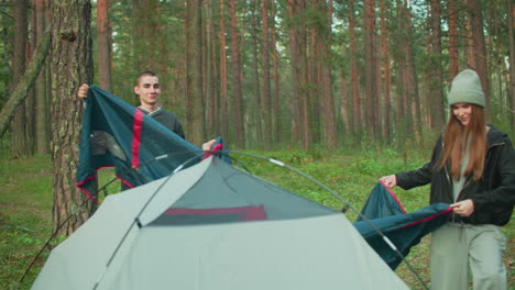 couple works together to cover camping tent with outer sheet in forest clearing, both smiling and adjusting material while surrounded by tall trees and natural greenery