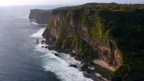 Aerial-view-of-majestic-cliffs-standing-tall-beside-coastline-of-beach-and-low-tides-moving-towards-them