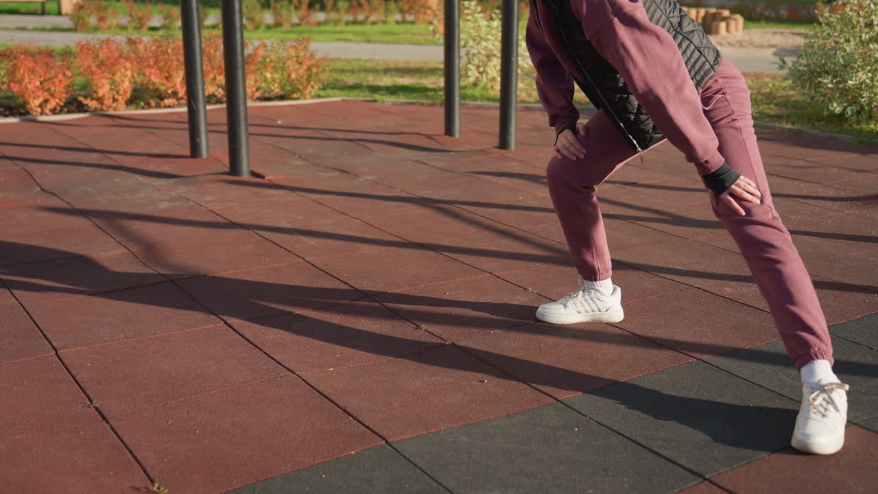 Female athlete under sunny weather performing side lunge workout with hands on legs on outdoor pavement in modern fitness park framed by flowers and greenery, long shadows across ground