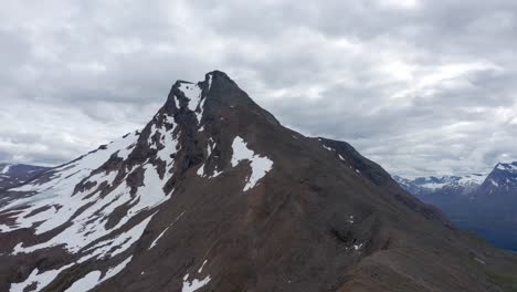 Drone-shot-of-dramatic-peak-in-northern-Norway-surrounded-by-snow-and-mountains-with-dramatic-sky