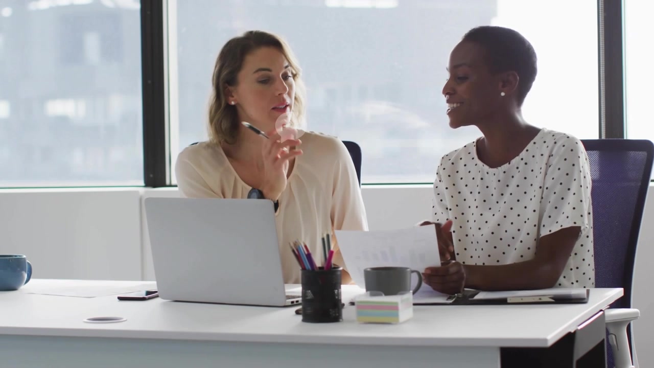 Two women at desk left woman gesturing making chat bubbles around laptop document business review