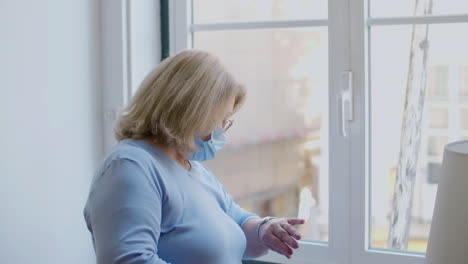 Side-view-of-mature-woman-in-medical-mask-standing-near-window