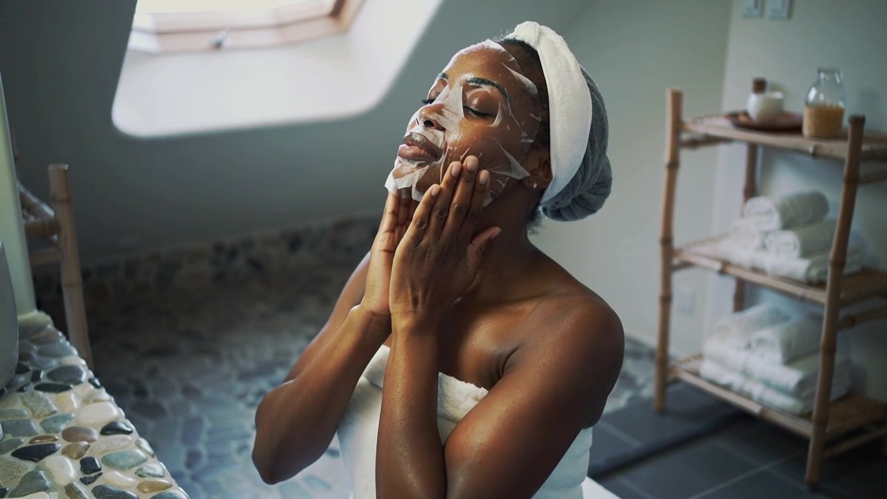 Applying woman starting skincare smoothing white sheet mask at vanity wearing white towel+headband