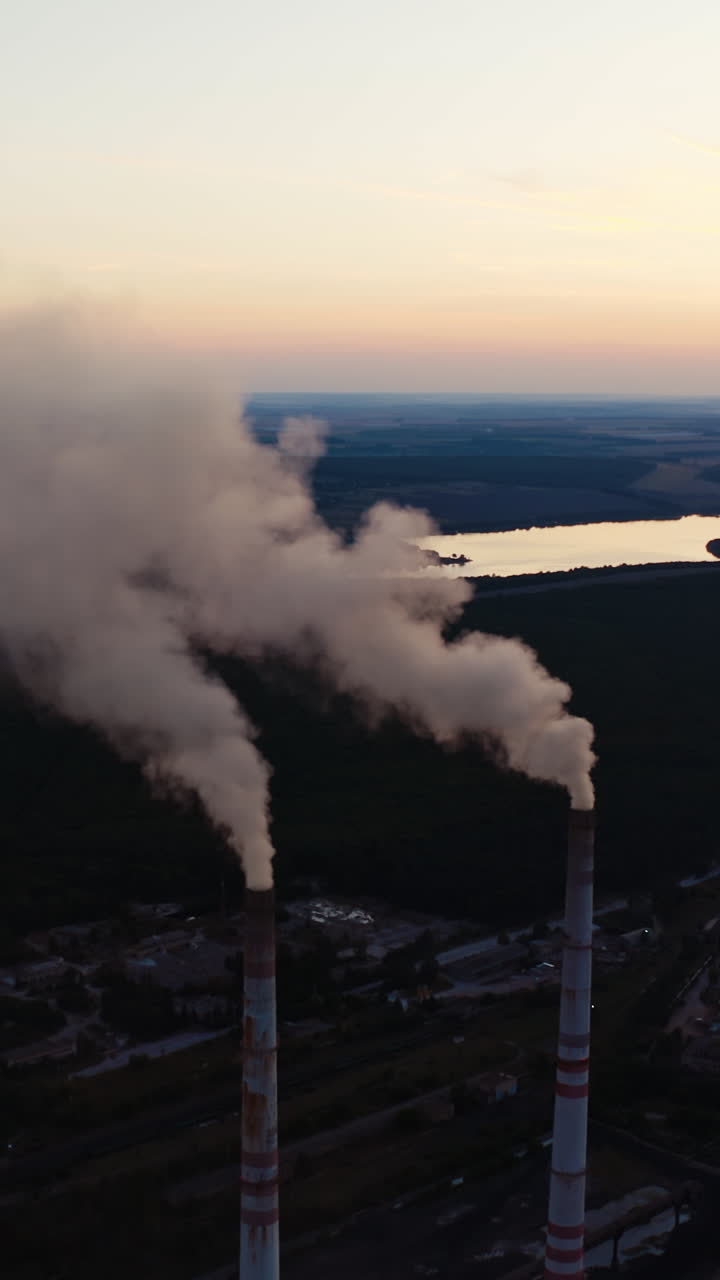 White smoke from pipes in the natural environment. Industrial factory near the beautiful river in the countryside in the evening. Air pollution. Vertical video