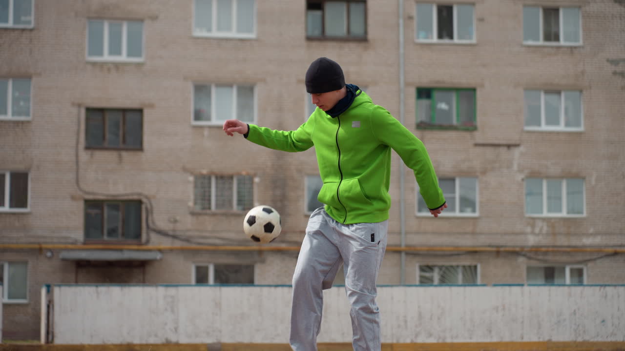 Hombre caucásico practicando fútbol freestyle en el patio de un apartamento, chaqueta verde y gorro, ejercicios de habilidad individuales sobre un fondo de hormigón, juego de pies concentrado y determinación urbana capturados en movimiento