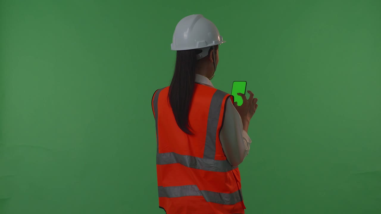 안전 헬을 입은 아시아 여성 엔지니어가 녹색 스크린 스마트폰에서 작업하고, 일몰 또는 일출 시간 동안 녹색 스클린 배경 스튜디오에 서있는 동안 주변을 둘러보고 있습니다.