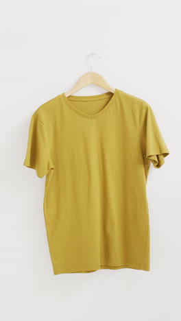 vertical video of close up of mustard t shirt hanging on white background with copy space
