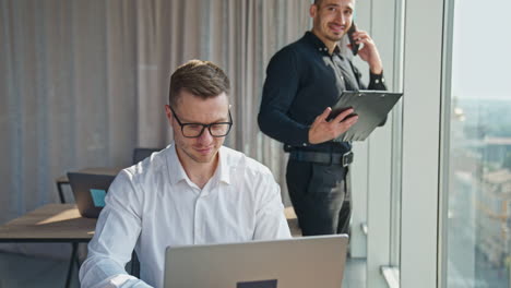 male business partners working in the office. man in black shirt has phone conversation and man in white shirt works on laptop.