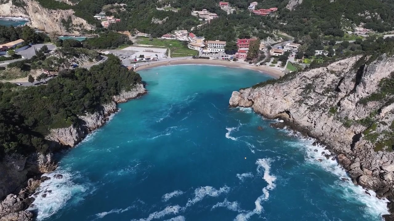 Volo di drone ad alta angolazione che entra nella suggestiva baia di Agios Spyridon a Paleokastritsa, Corfù. Onde del mare turchesi si riversano su una spiaggia sabbiosa circondata da verdi colline.