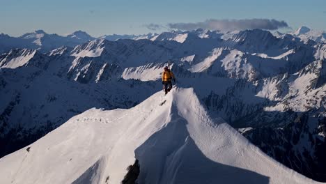 Un-Escalador-Se-Encuentra-En-La-Cima-De-Una-Montaña-Nevada-En-Los-Confines-Salvajes-De-La-Naturaleza-Con-Enormes-Montañas-Y-Valles-Gigantes-Al-Fondo