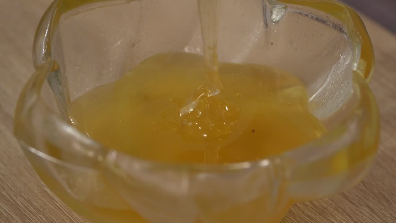Golden honey drips into a clear glass bowl, forming a smooth, thick pool with a glossy texture and rich amber tone on a wooden surface, emphasizing natural sweetness and viscous movement in close up