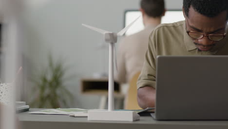 hombre de negocios que usa una computadora portátil sentada en la mesa con un modelo de molino de viento en la oficina 1