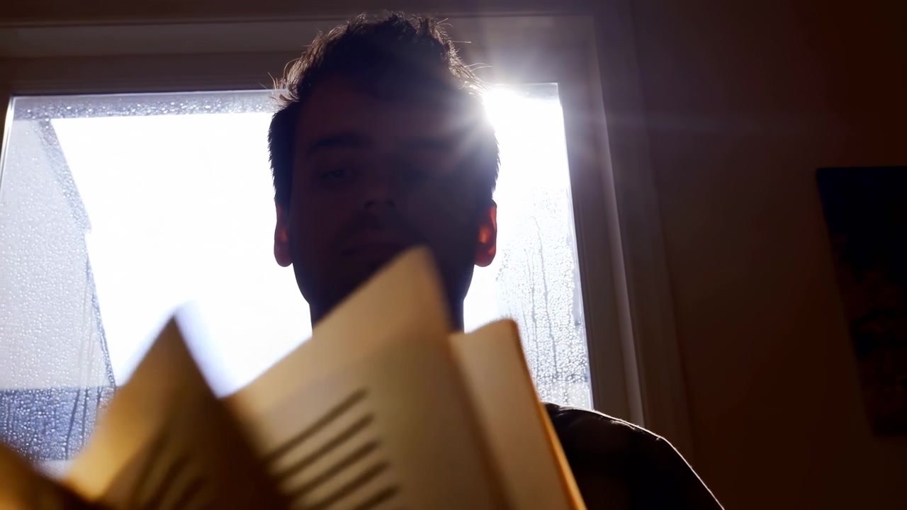 Man looking out the window, in interior room with large windows, with window, wall, from a over-the-shoulder shot from behind the subject perspective, with yellow, brown accents, nearby objects.