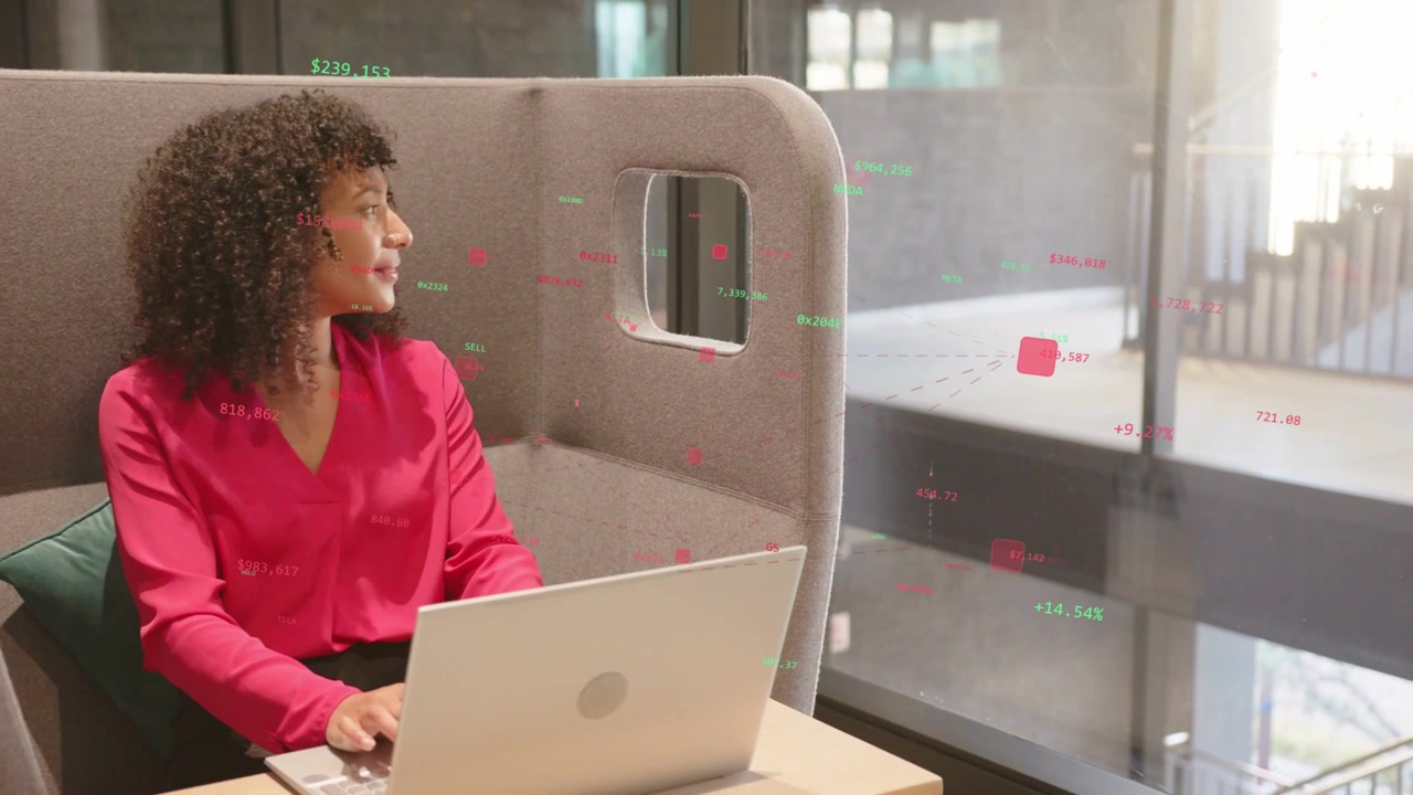 Woman in grey booth typing on laptop, reviewing data, causing red-green numbers reflecting on glass