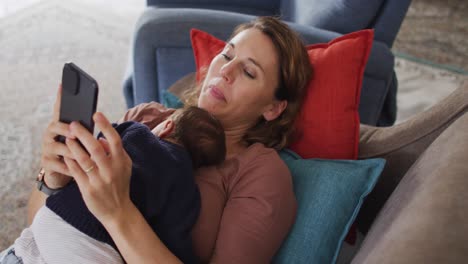 Video-of-caucasian-mother-with-smartphone-lying-on-sofa-with-newborn-sleeping-baby