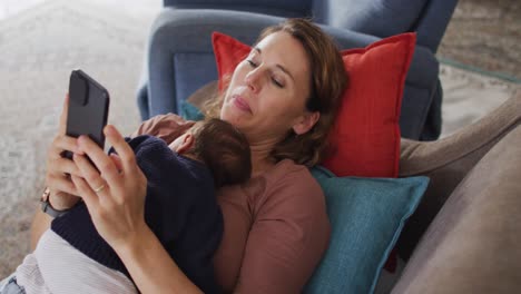 video of caucasian mother with smartphone lying on sofa with newborn sleeping baby