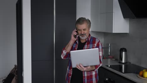 Anciano-Barbudo-Usando-Una-Laptop-En-La-Cocina-Y-Hablando-Por-Teléfono-Al-Mismo-Tiempo