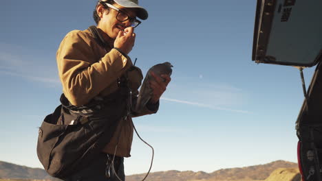 low medium shot of a man tying a pigeon on a leash in preparation to train a bird for falconry as a lure smiling in front of farm fields with mountains in the distance