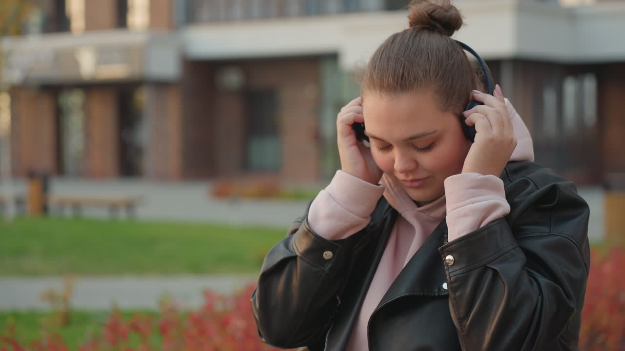 Woman adjusting headphone while seated outdoors enjoying peaceful morning breeze with relaxed expression in soft lighting, surrounded by red plants and slightly blurred background