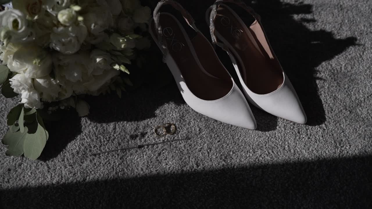 White bridal shoes and bouquet on a carpet with wedding rings in soft sunlight.