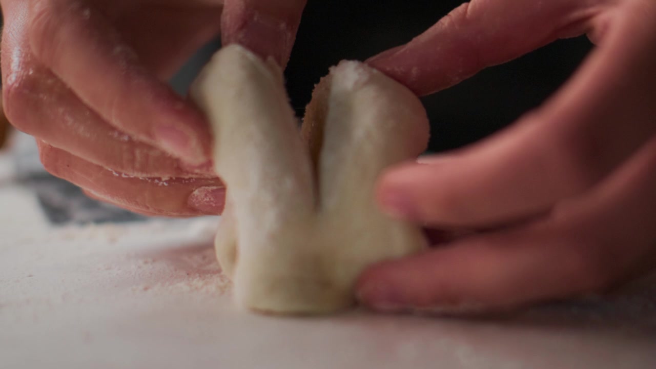Mani modellano delicatamente l'impasto su una superficie infarinata. Le dita premono e piegano, creando una consistenza liscia. Un primo piano evidenzia il processo tattile e culinario. Ideale per contenuti di cucina, panificazione o preparazione alimentare.