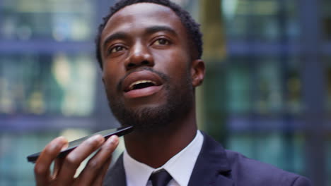 Close-Up-Of-Young-Businessman-Wearing-Suit-Talking-On-Mobile-Phone-Using-Built-In-Microphone-Standing-Outside-Offices-In-The-Financial-District-Of-The-City-Of-London-UK-Shot-In-Real-Time