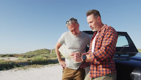 Happy-caucasian-gay-male-couple-leaning-on-car,-reading-map-and-talking-on-sunny-day-at-the-beach