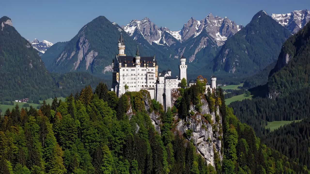 Aerial view of a fairytale castle atop a forested hill, surrounded by majestic mountains