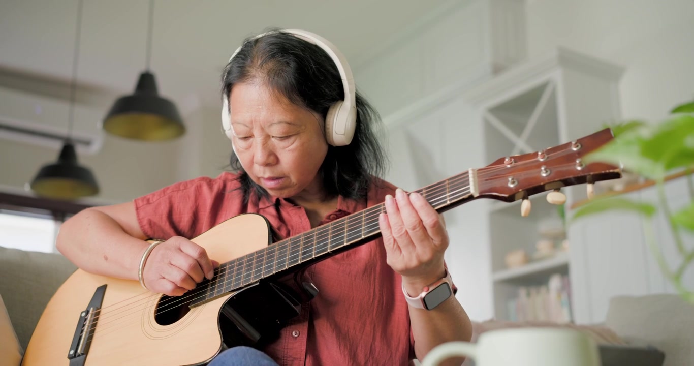 Donna anziana asiatica che segue una lezione su tablet, indossando cuffie, accordando e suonando la chitarra sul divano.