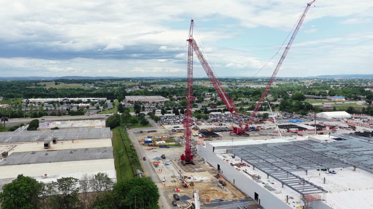 Twin red cranes tower above data center under construction, with steel frame and rolling Pennsylvania farmland under partly cloudy skies in Lancaster, Pennsylvania, USA. Aerial drone wide shot.