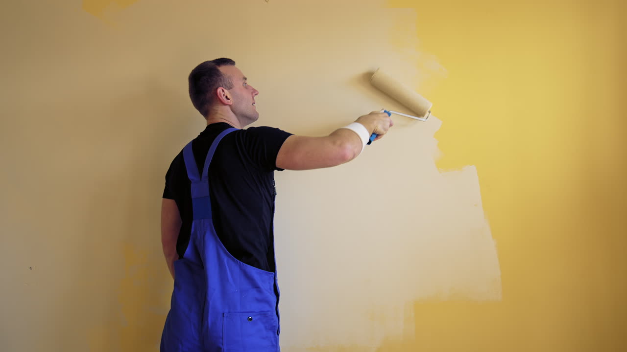 Painter renewal room interior with a paint roller. Young builder is painting wall in the room. Worker changing color into lightsome in the apartment. Rear view.