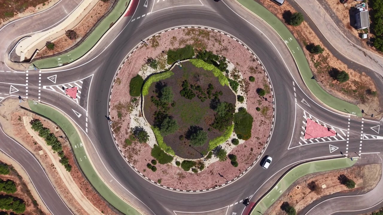 Top-down view of a road roundabout with a circular landscaped island featuring shrubs and trees, surrounded by asphalt lanes with road markings and two cars