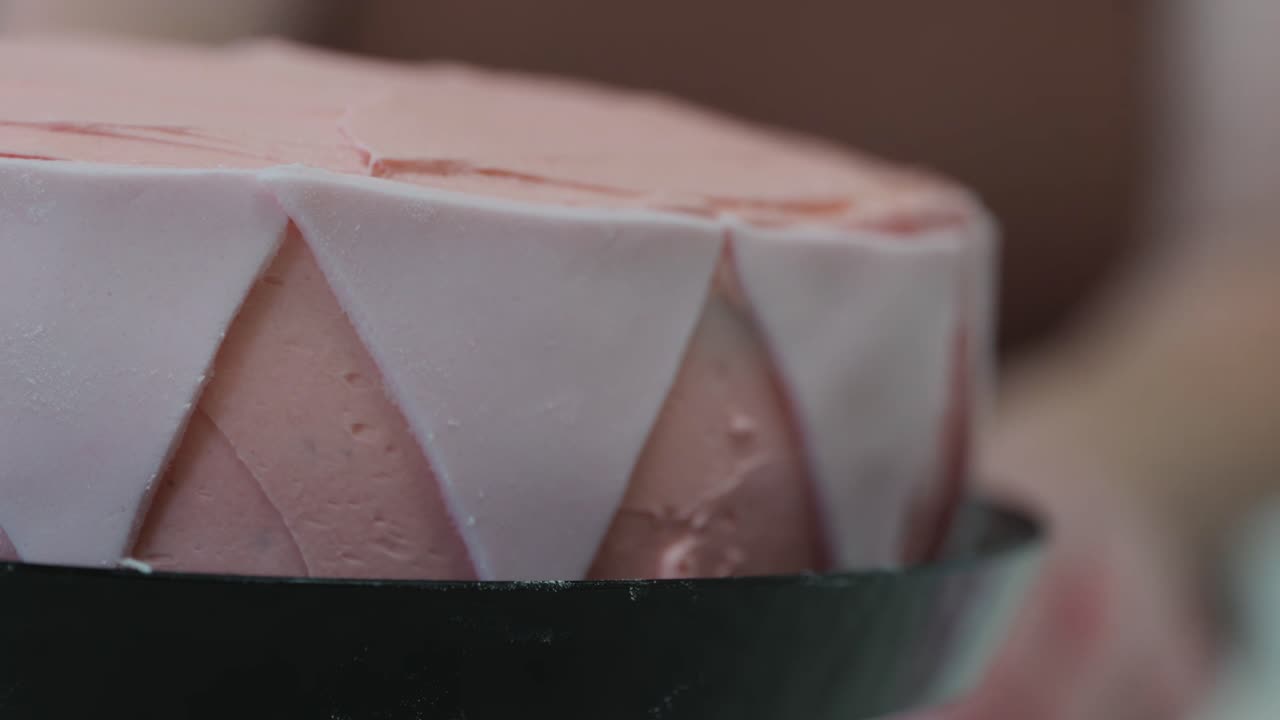 A person places pink fondant triangles onto the edge of a birthday cake decorated with pink buttercream frosting