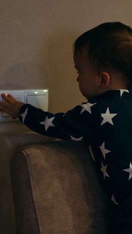 dark-haired caucasian toddler standing on the sofa. baby boy reaches his hand to the switch and turns the light on and off. vertical video