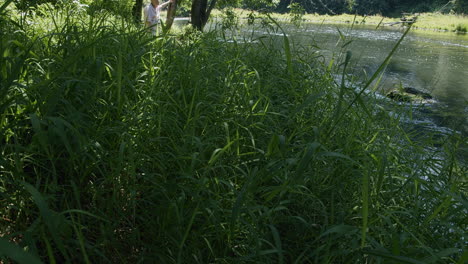 older-man-leans-on-tree-in-tall-grass-by-slow-moving-river,-tilt-up