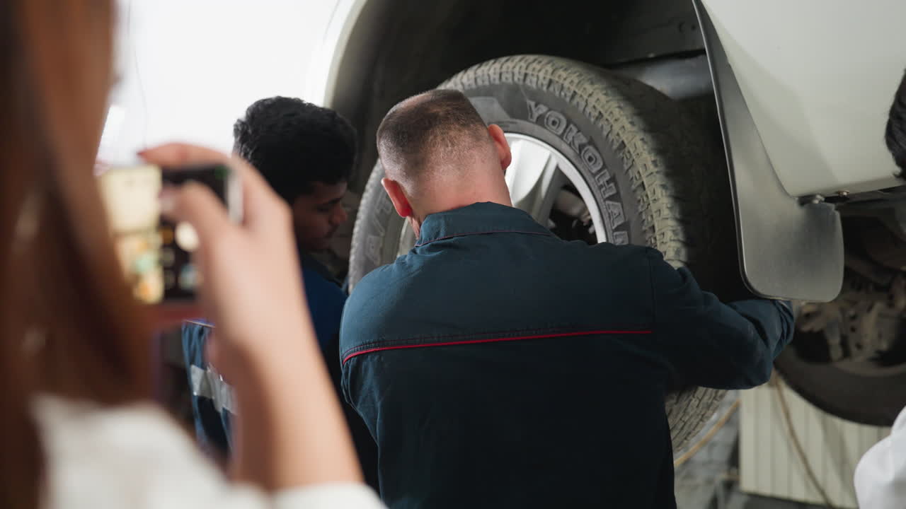 vista de fondo de un inventor en bata de laboratorio fotografiando a un mecánico arreglando un neumático de coche con un teléfono inteligente mientras un estudiante mira, con los brazos cruzados, en un taller mecánico