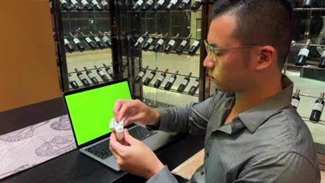 asian millennial male looking at laptop green screen, wearing earbuds, and enjoying music in luxurious home