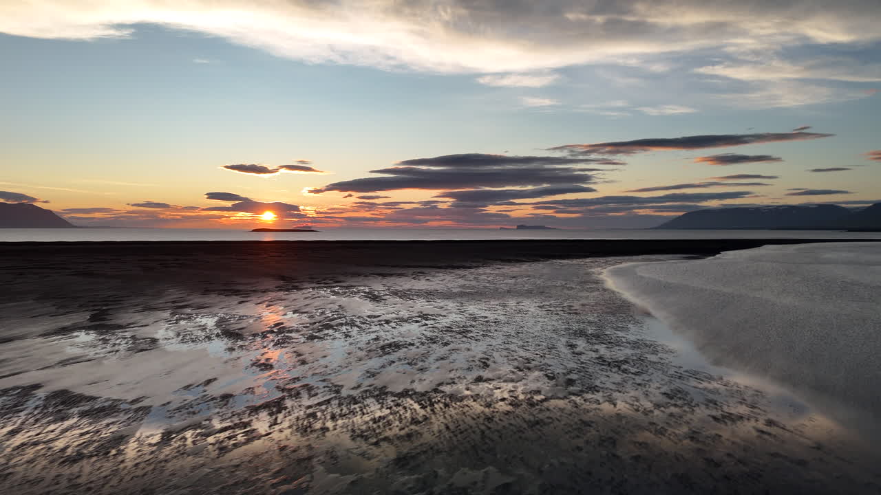 Fredfyldt solnedgang over en øde, sort sandstrand ved Skagafjörður-fjorden nær Sauðárkrókur i Nordisland med roligt havvand, fjerne bjerge og blødt, gyldent lys, der reflekteres i horisonten